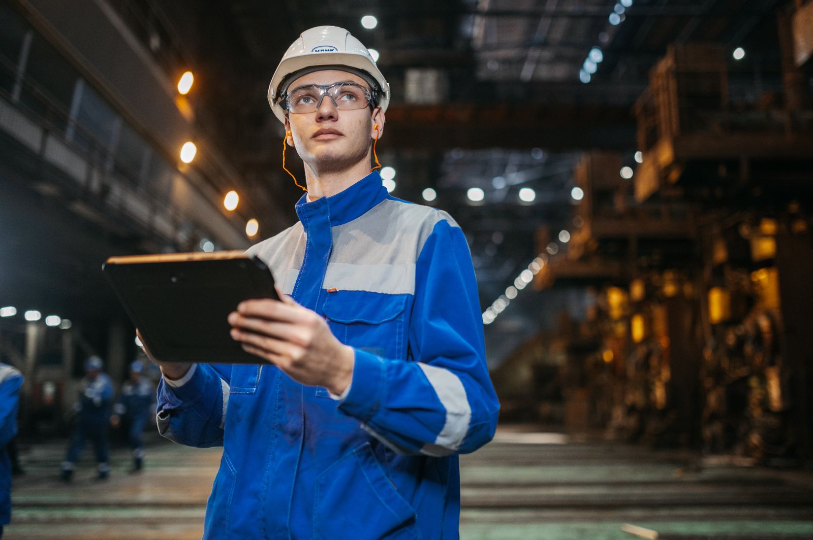 A worker in safety gear reading a tablet on a factory floor