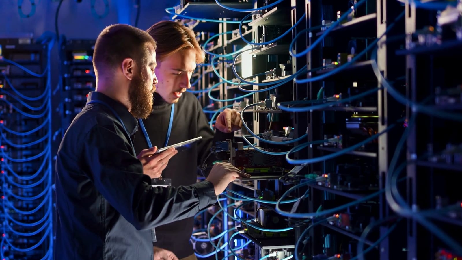 Two technicians stand at an open server rack in a data hall; one holds a tablet while the other works on the equipment