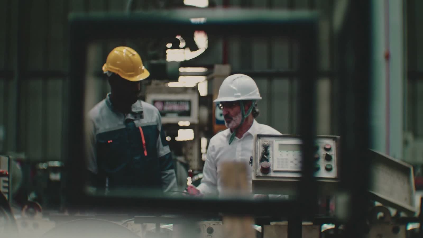 A senior mechanic in a white hard hat talks a younger colleague through a machine on a factory floor